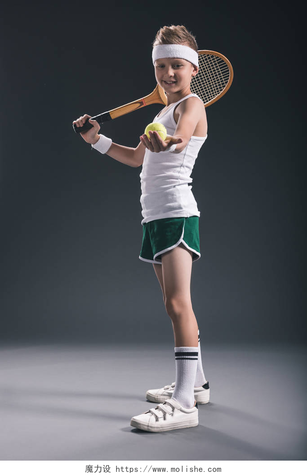 在灰色的背景墙和地面上一个穿着白色上衣的网球男孩带网球拍和球在黑暗背景的运动服的微笑男孩